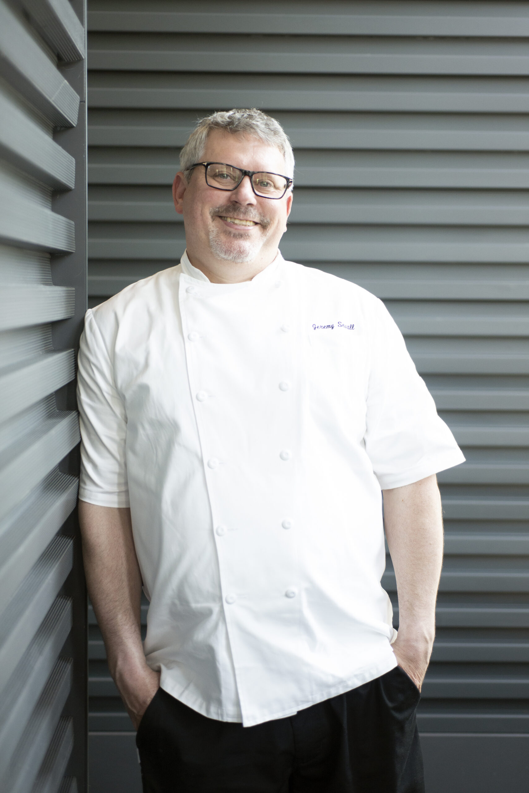 Man wearing chef's coat and leaning against a wall.